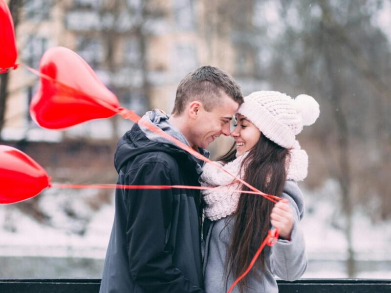 Young couple embracing with red heart balloons outdoors in winter, sharing a joyful moment.