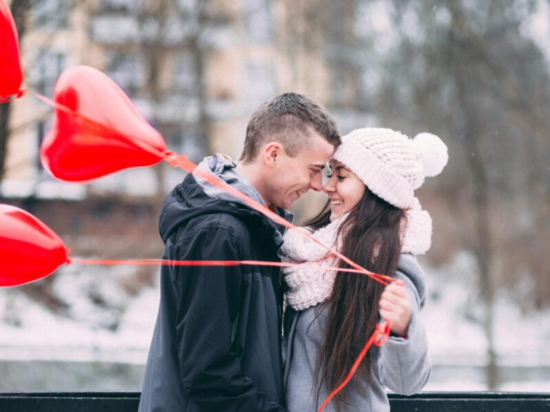Young couple embracing with red heart balloons outdoors in winter, sharing a joyful moment.
