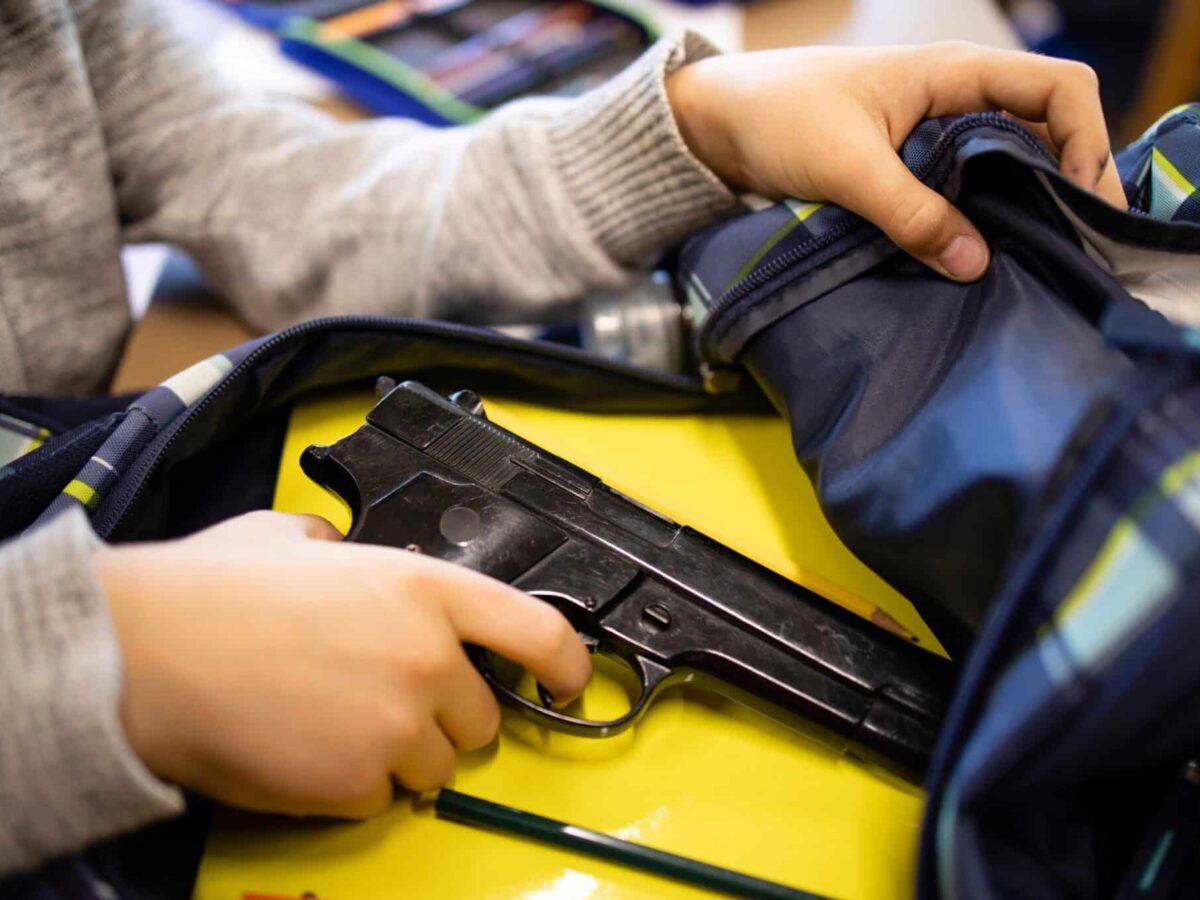 Child's hands holding a black handgun inside a backpack on a yellow table.