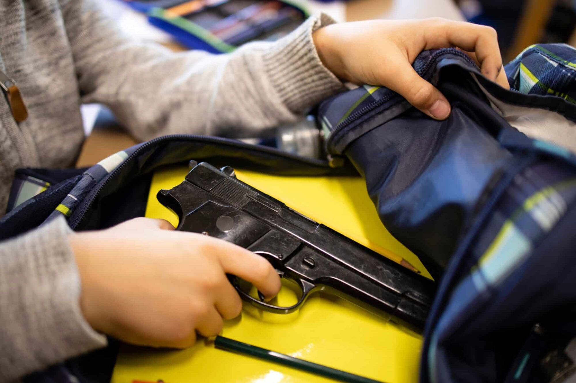 Child's hands holding a black handgun inside a backpack on a yellow table.