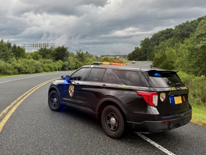 Police SUV with flashing lights blocking a wet rural highway under dark storm clouds.