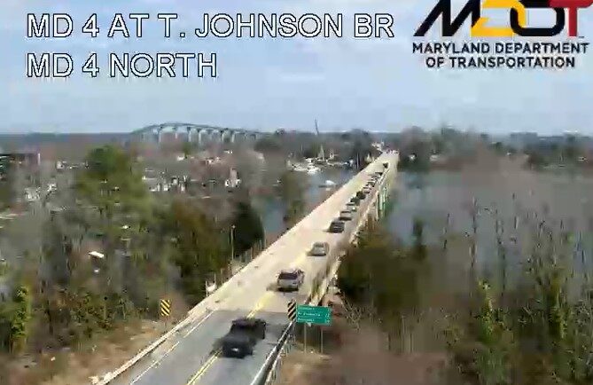 Traffic crosses a long bridge over a river with trees and a distant truss bridge.