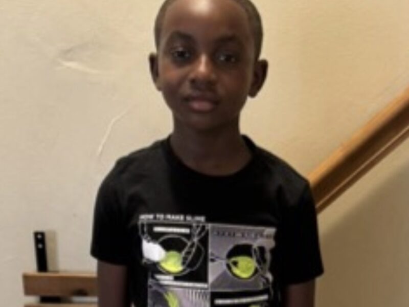 Young boy standing indoors by a staircase, wearing a black graphic T-shirt.