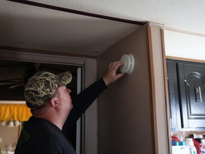 Man in a camouflage cap installing a smoke detector above a doorway indoors.