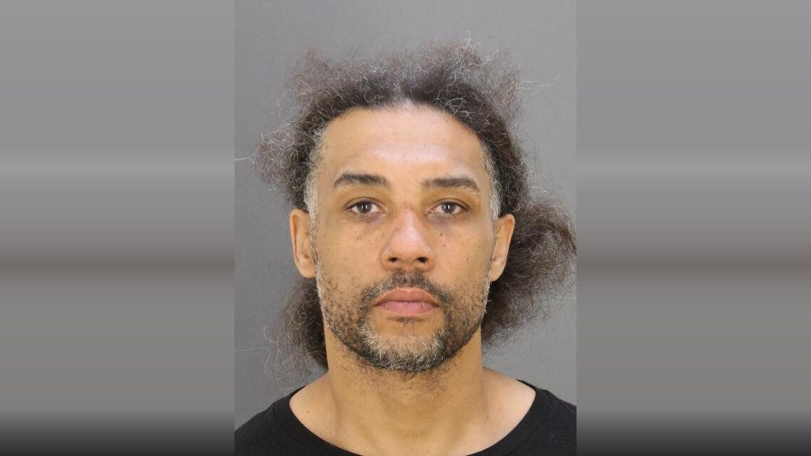 Mugshot-style portrait of a man with curly hair and a neutral expression against a gray background.