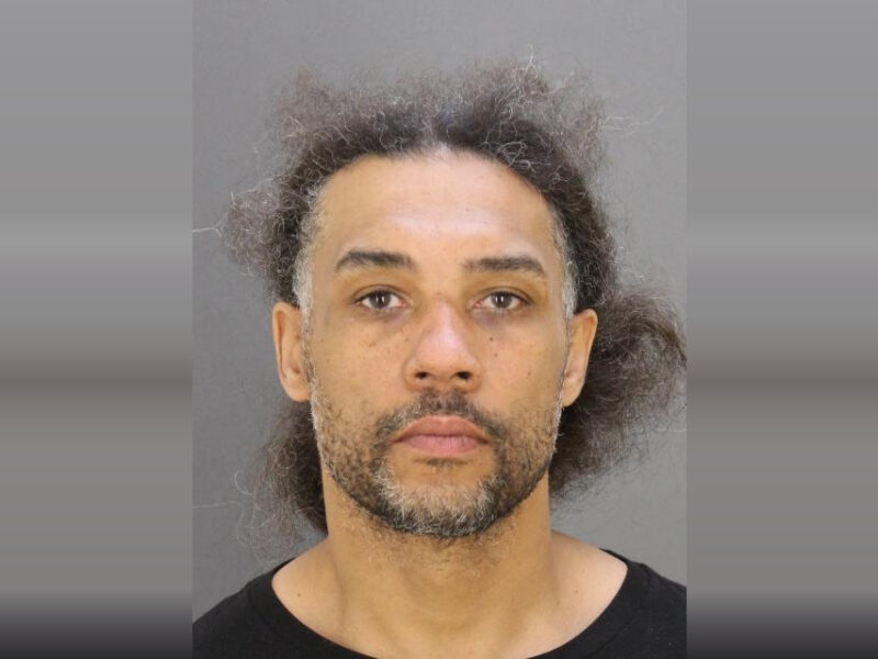 Mugshot-style portrait of a man with curly hair and a neutral expression against a gray background.