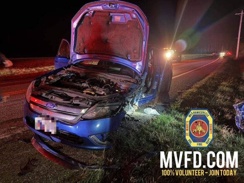 Blue car with hood open after a roadside crash at night, illuminated by emergency lights.