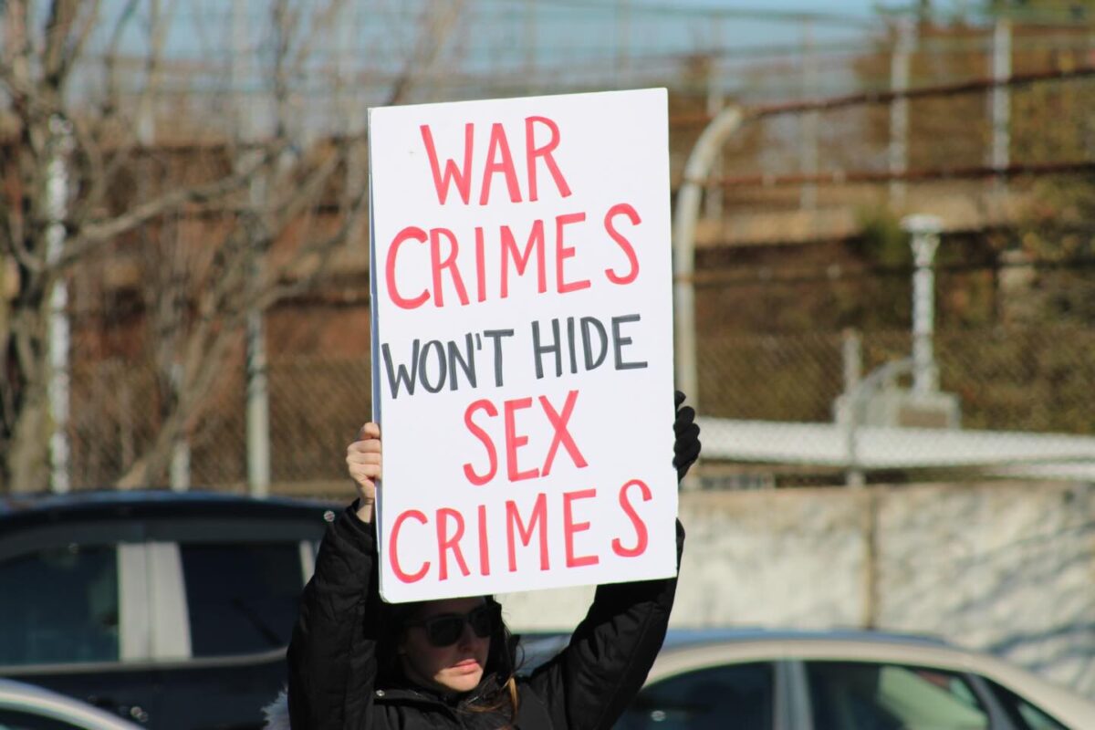 Protester holds a sign reading "WAR CRIMES WON'T HIDE SEX CRIMES" outdoors.