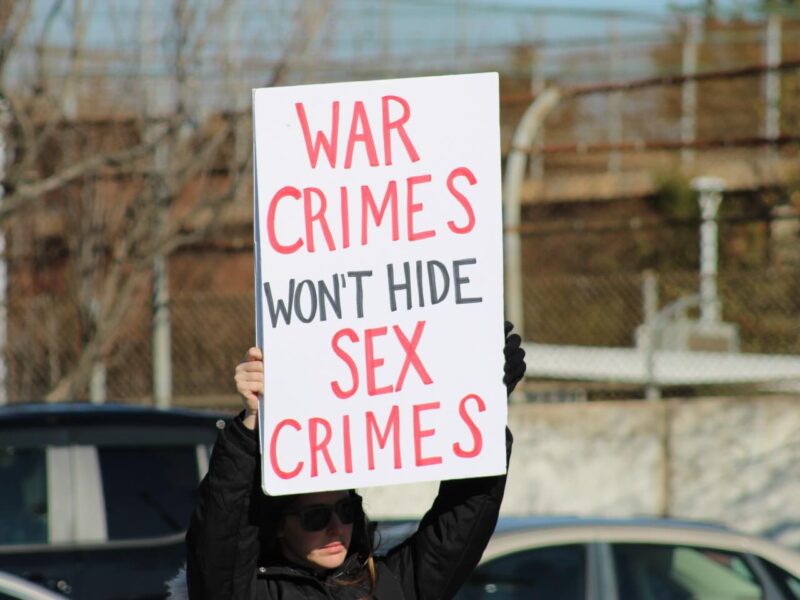 Protester holds a sign reading "WAR CRIMES WON'T HIDE SEX CRIMES" outdoors.