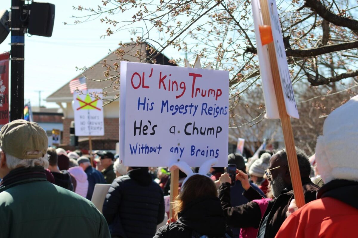 Crowd at a protest holds a sign mocking Trump with handwritten text.