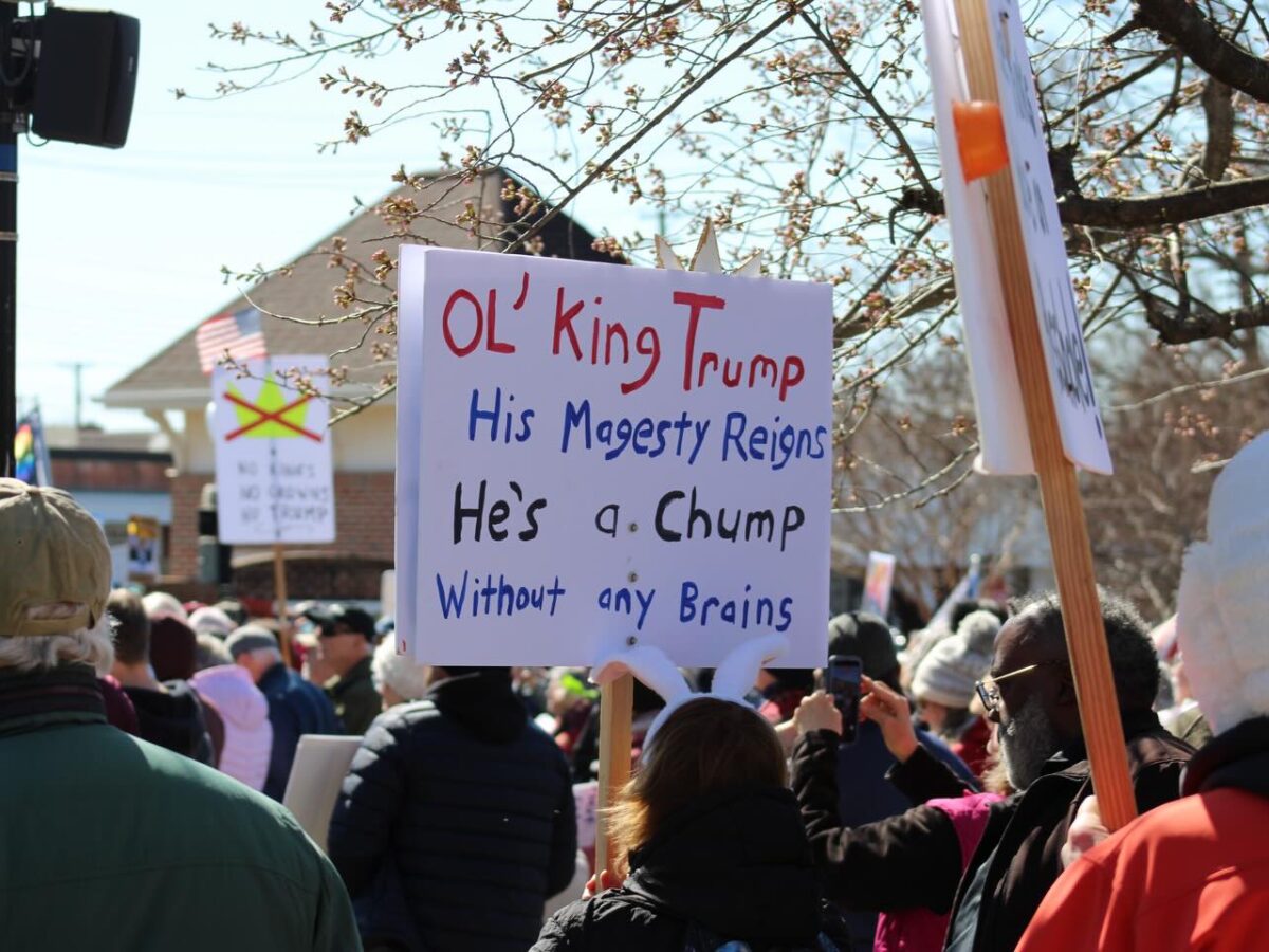 Crowd at a protest holds a sign mocking Trump with handwritten text.