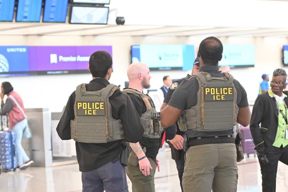 Three ICE police officers wearing tactical vests with "POLICE ICE" patches on their backs are standing and talking in an airport terminal. In the background, there are flight information screens and a few travelers, including a woman with a suitcase and a person in a black and green uniform.