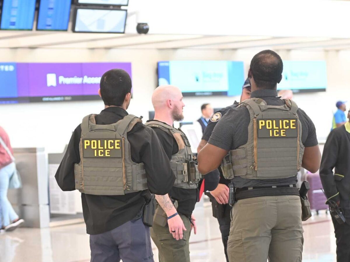 Three ICE police officers wearing tactical vests with "POLICE ICE" patches on their backs are standing and talking in an airport terminal. In the background, there are flight information screens and a few travelers, including a woman with a suitcase and a person in a black and green uniform.