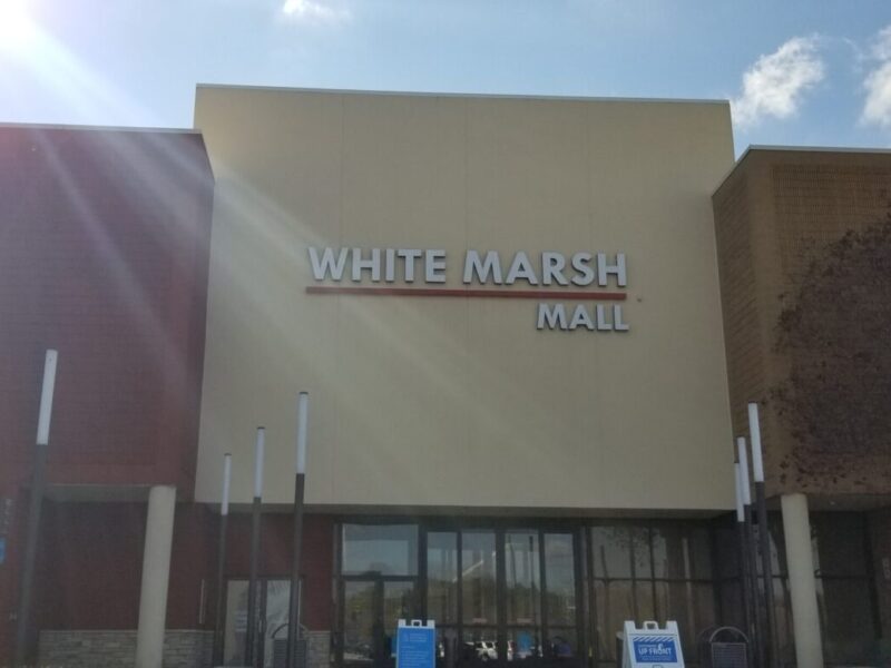 Exterior of White Marsh Mall under a bright blue sky with sun glare.