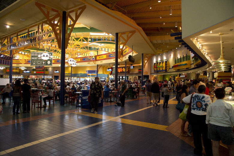Crowded indoor food court with shoppers, tables, and bright decorative lighting.