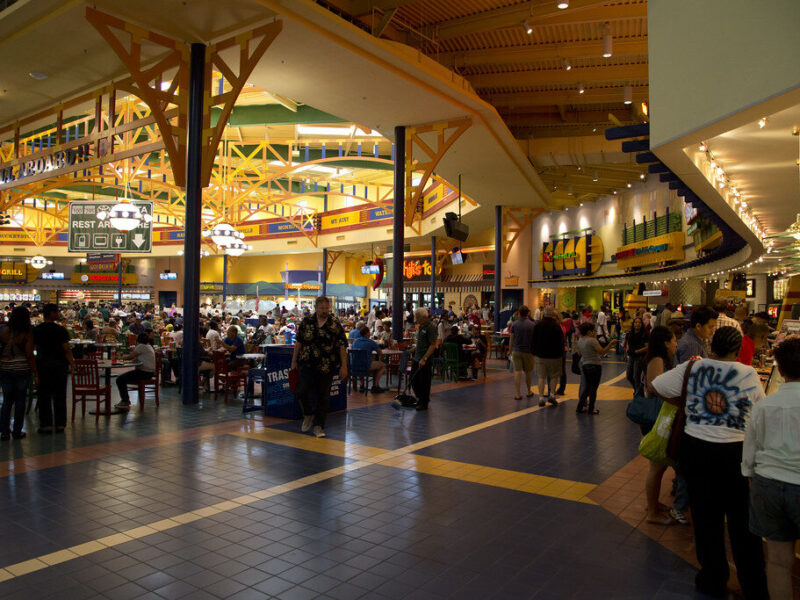 Crowded indoor food court with shoppers, tables, and bright decorative lighting.
