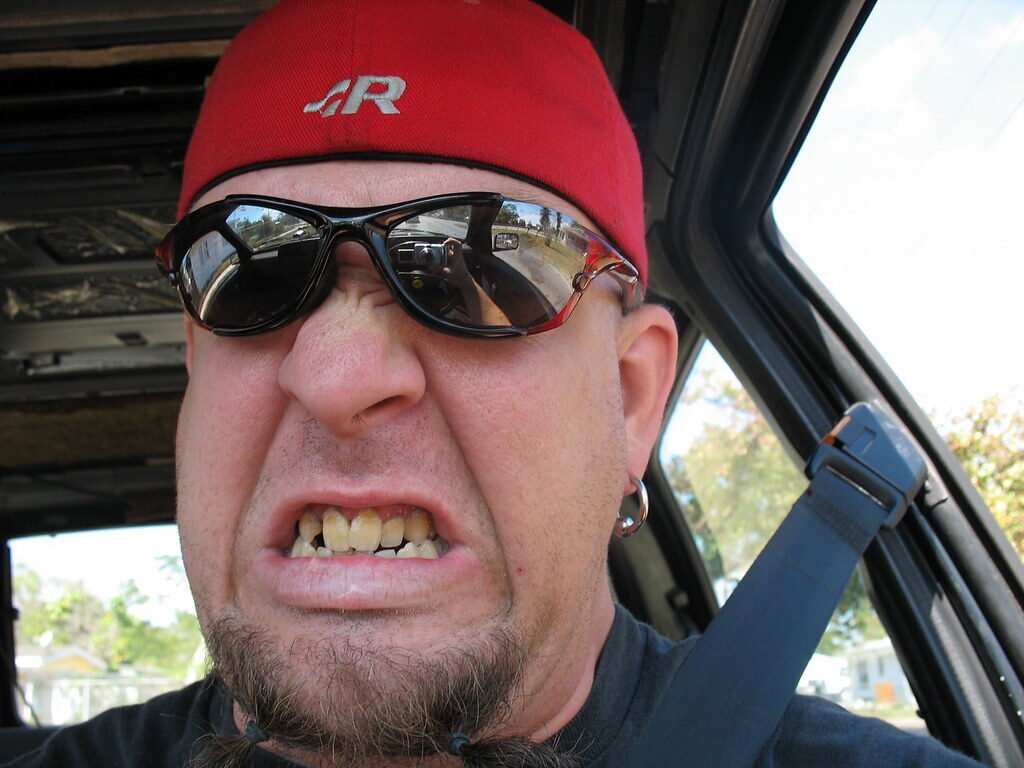 Man in red cap and sunglasses making a grimacing face inside a car.