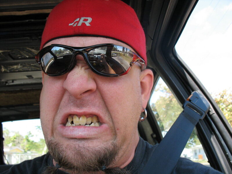 Man in red cap and sunglasses making a grimacing face inside a car.