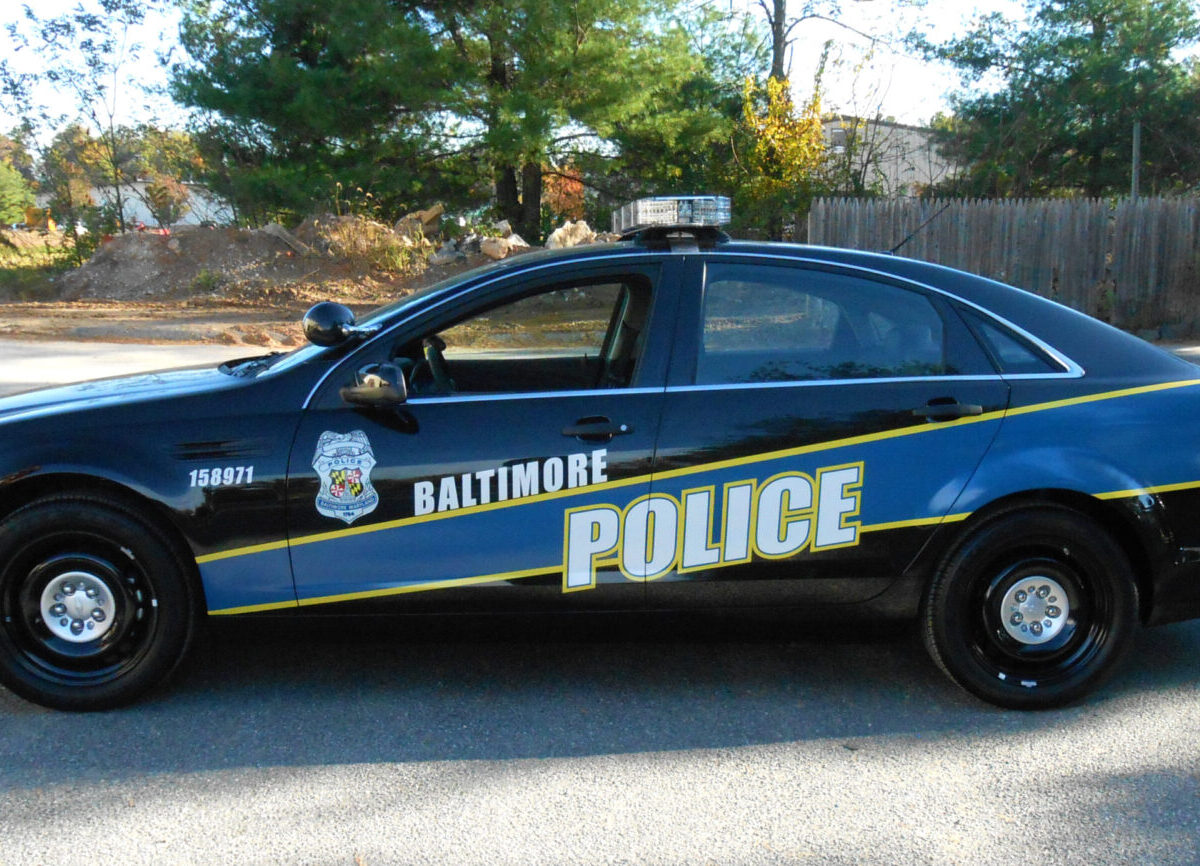 Baltimore police car parked on a road beside trees and a wooden fence.