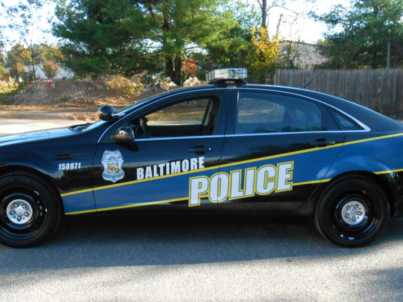 Baltimore police car parked on a road beside trees and a wooden fence.