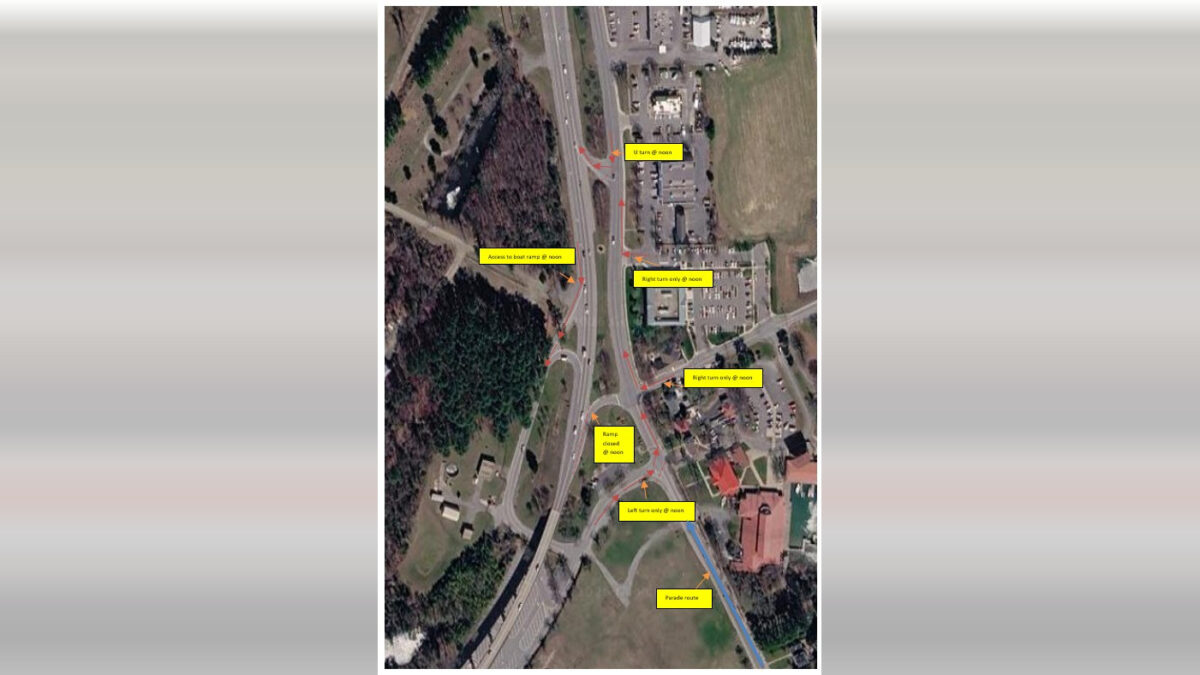 Satellite view of a highway interchange beside wooded land, buildings, and parking lots with yellow labels overlaid.