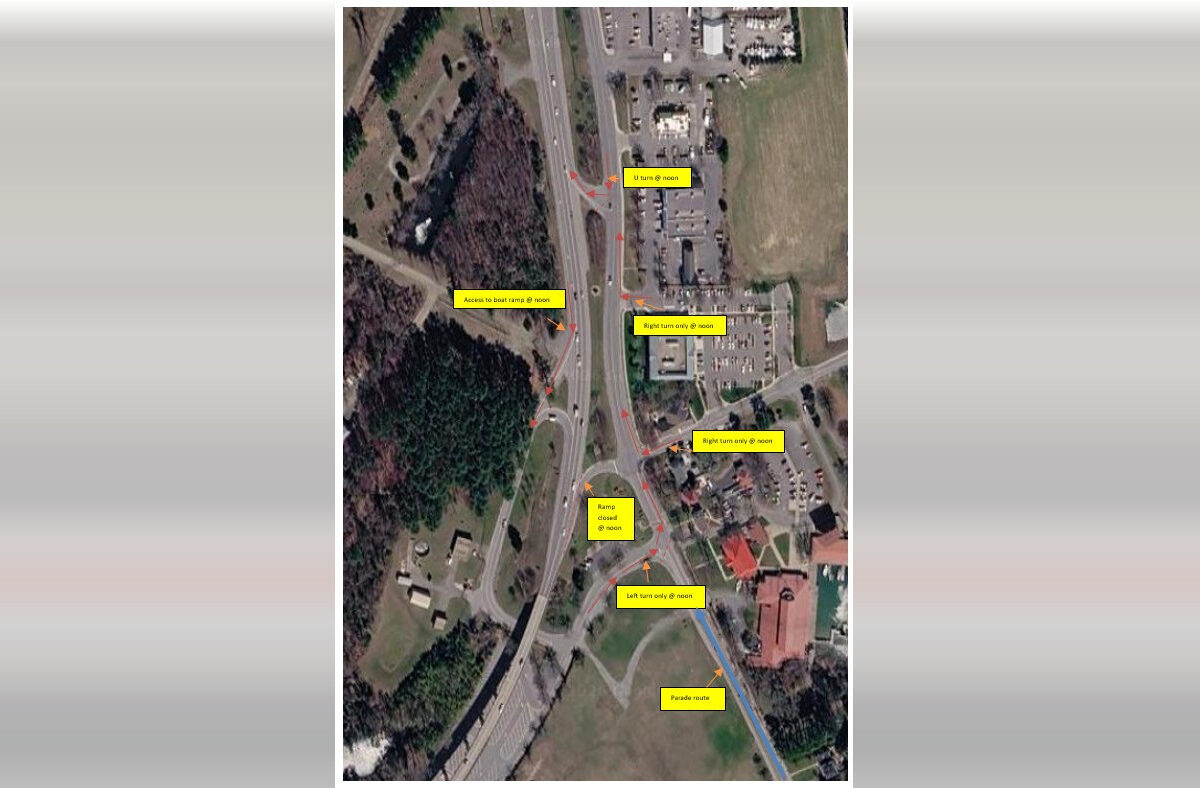 Satellite view of a highway interchange beside wooded land, buildings, and parking lots with yellow labels overlaid.