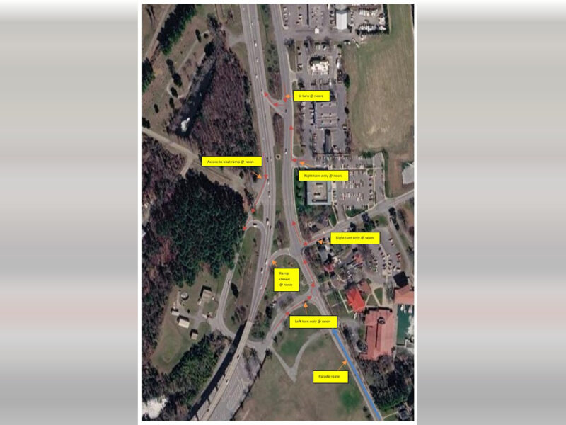 Satellite view of a highway interchange beside wooded land, buildings, and parking lots with yellow labels overlaid.