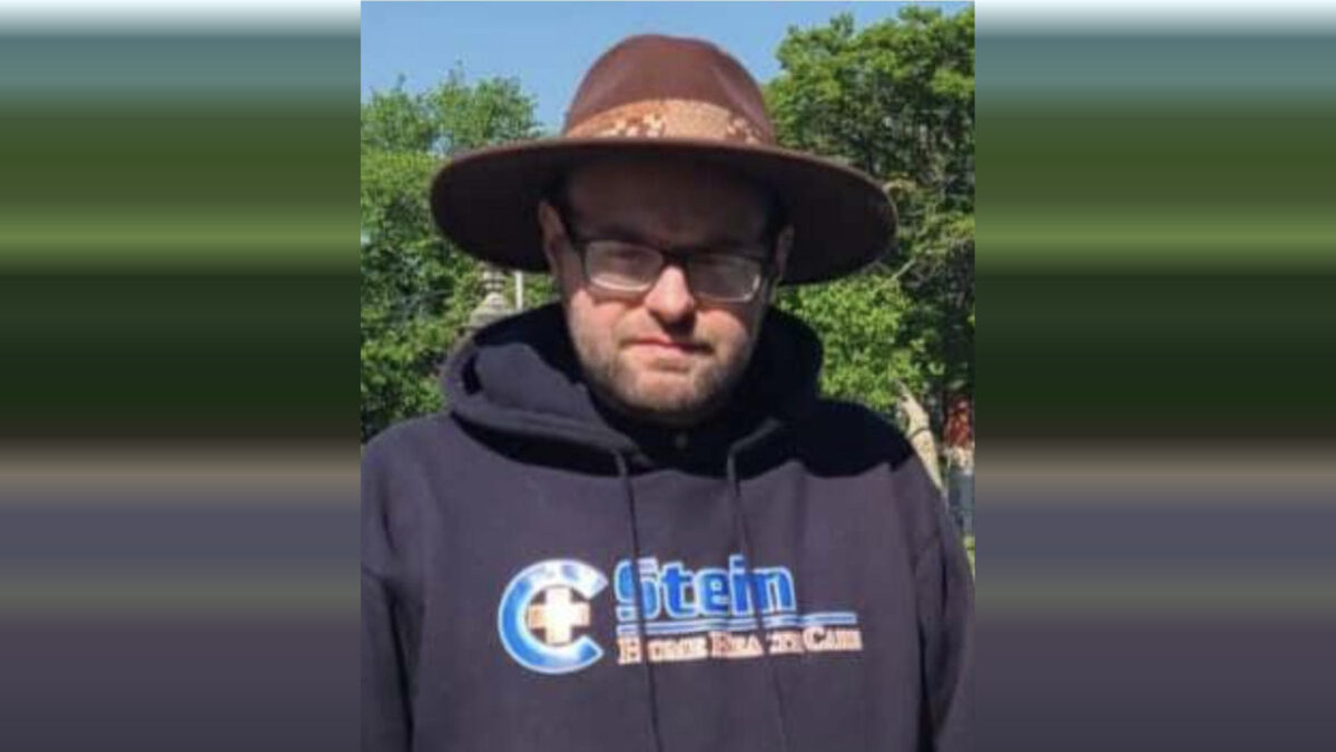 Man in a wide-brimmed hat and black hoodie standing outdoors in front of trees