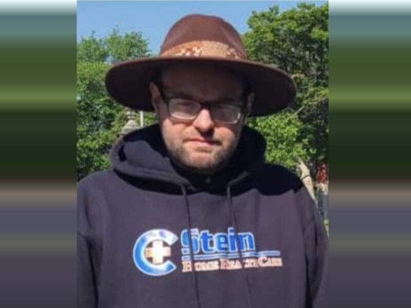 Man in a wide-brimmed hat and black hoodie standing outdoors in front of trees