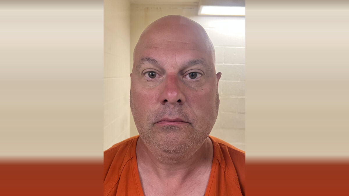 Bald man in an orange shirt faces the camera against a tiled bathroom background.