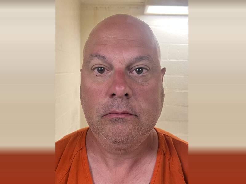 Bald man in an orange shirt faces the camera against a tiled bathroom background.