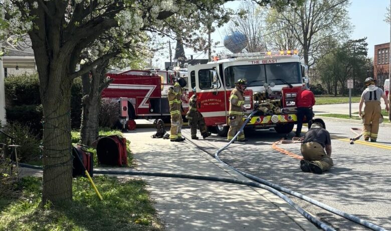 Firefighters work around a red fire truck at a roadside scene with hoses laid across the street.