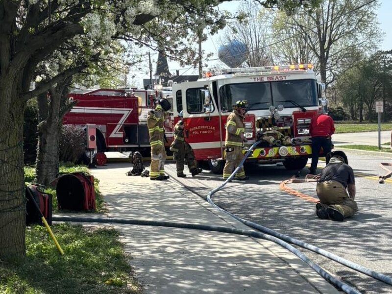 Firefighters work around a red fire truck at a roadside scene with hoses laid across the street.