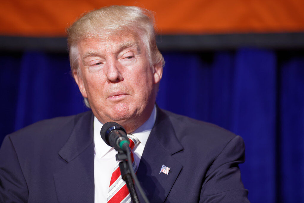 Donald Trump speaking at a podium with a microphone against a blue curtain backdrop.