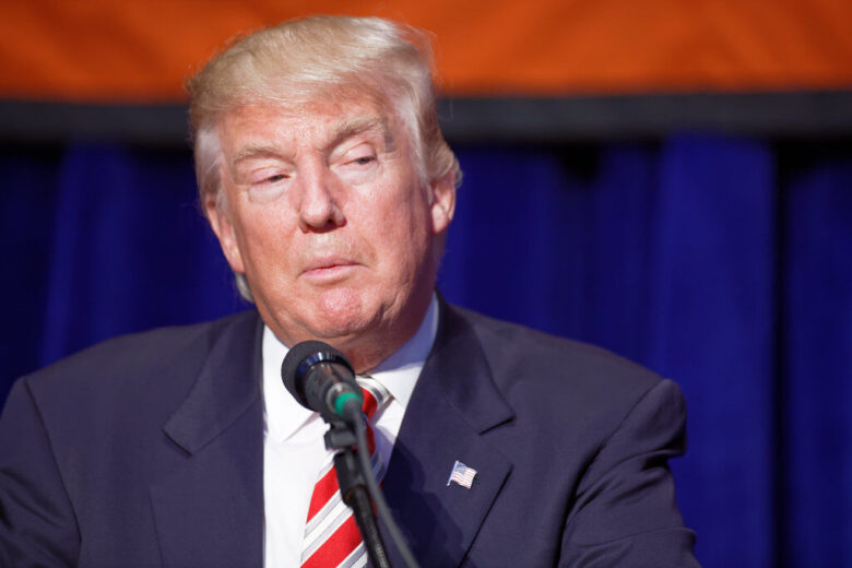 Donald Trump speaking at a podium with a microphone against a blue curtain backdrop.