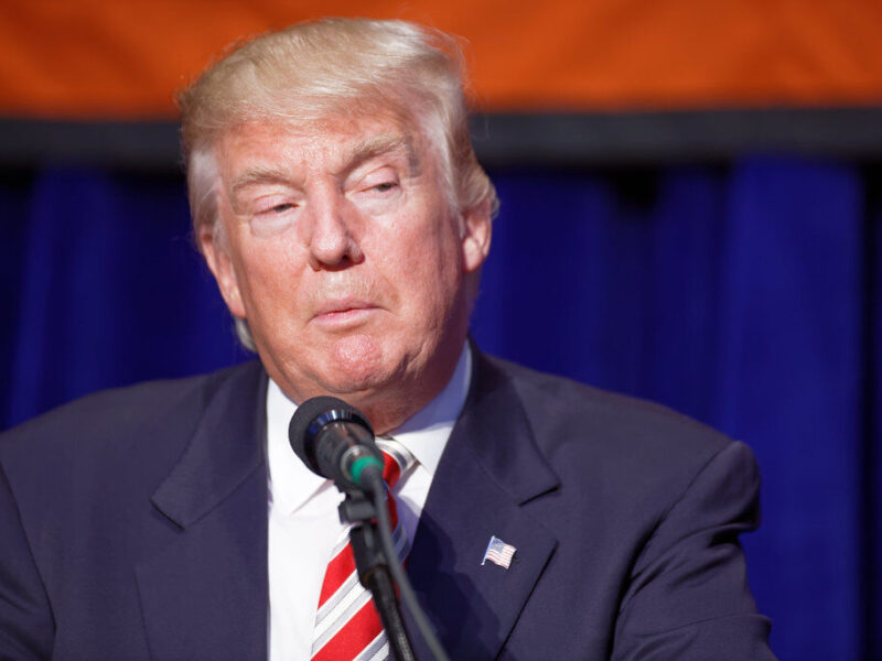 Donald Trump speaking at a podium with a microphone against a blue curtain backdrop.