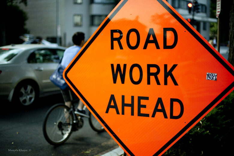 Bright orange road work ahead sign beside a cyclist on a city street.
