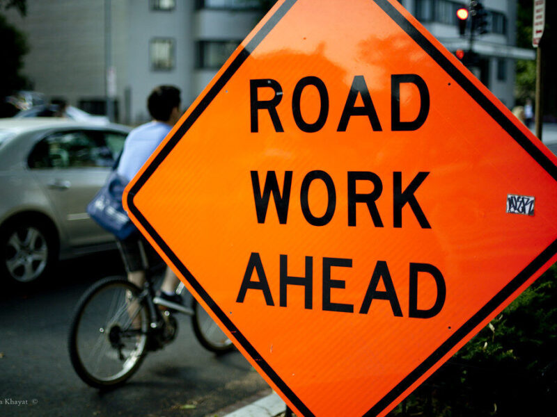 Bright orange road work ahead sign beside a cyclist on a city street.