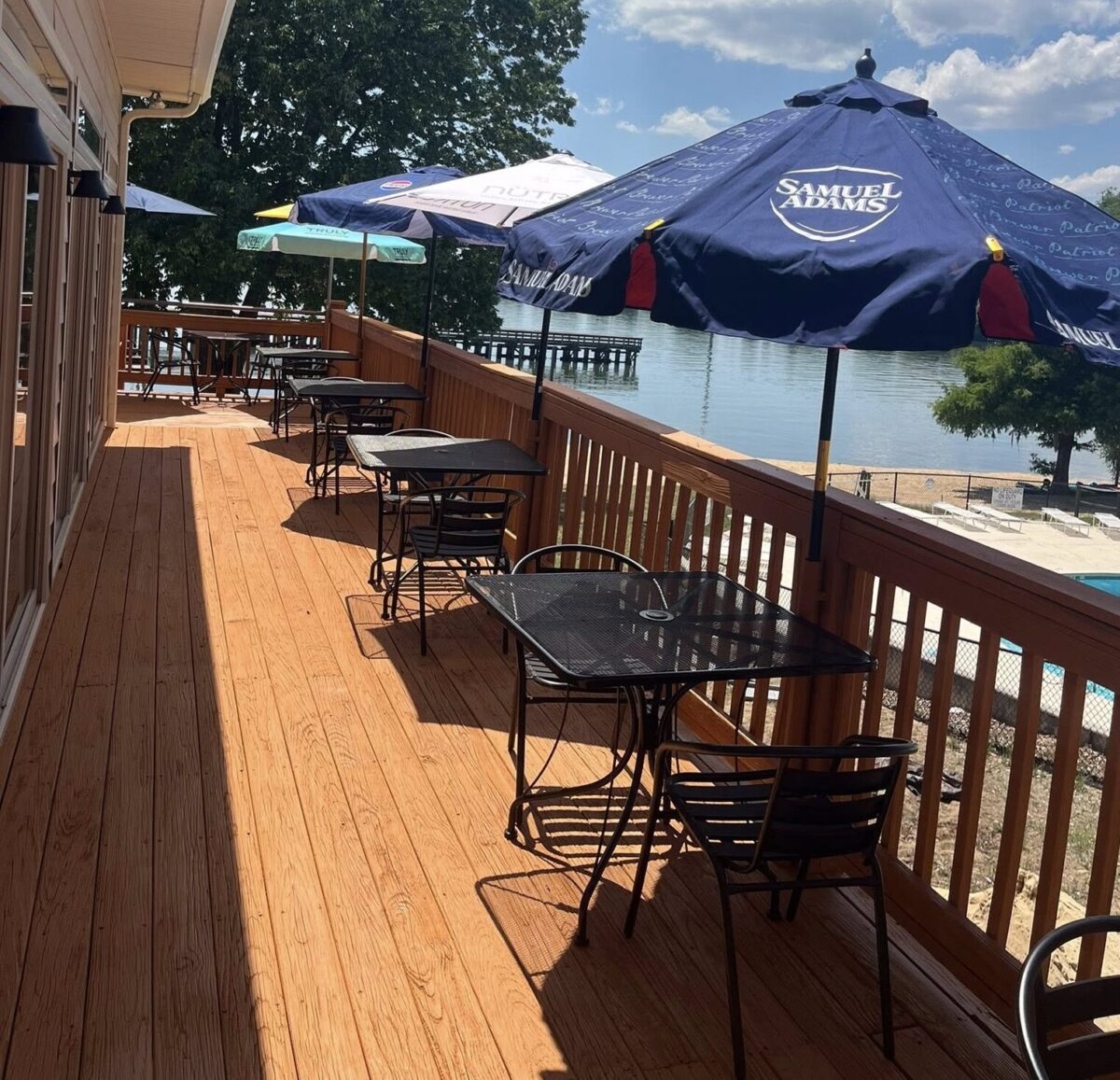 Wooden waterfront deck with empty tables, chairs, and blue umbrellas overlooking calm water.