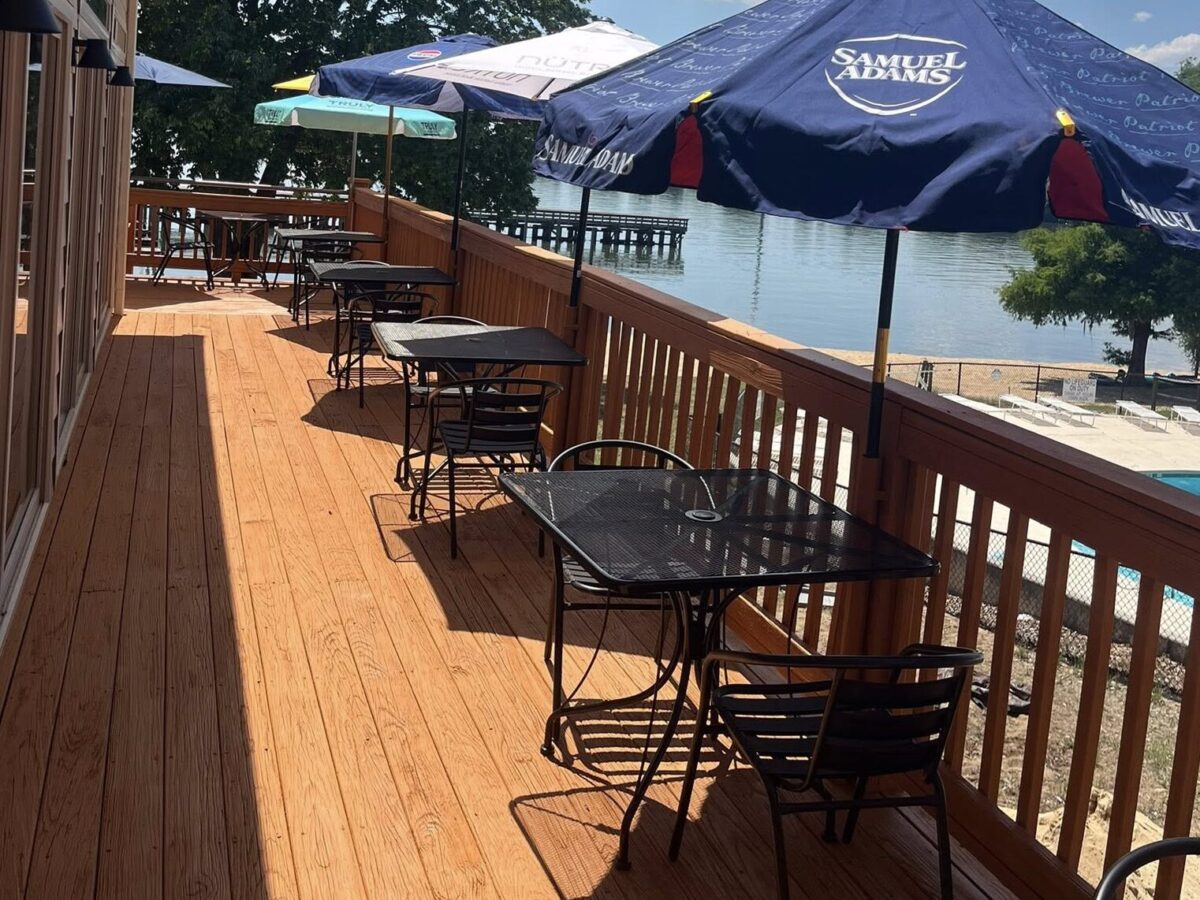 Wooden waterfront deck with empty tables, chairs, and blue umbrellas overlooking calm water.