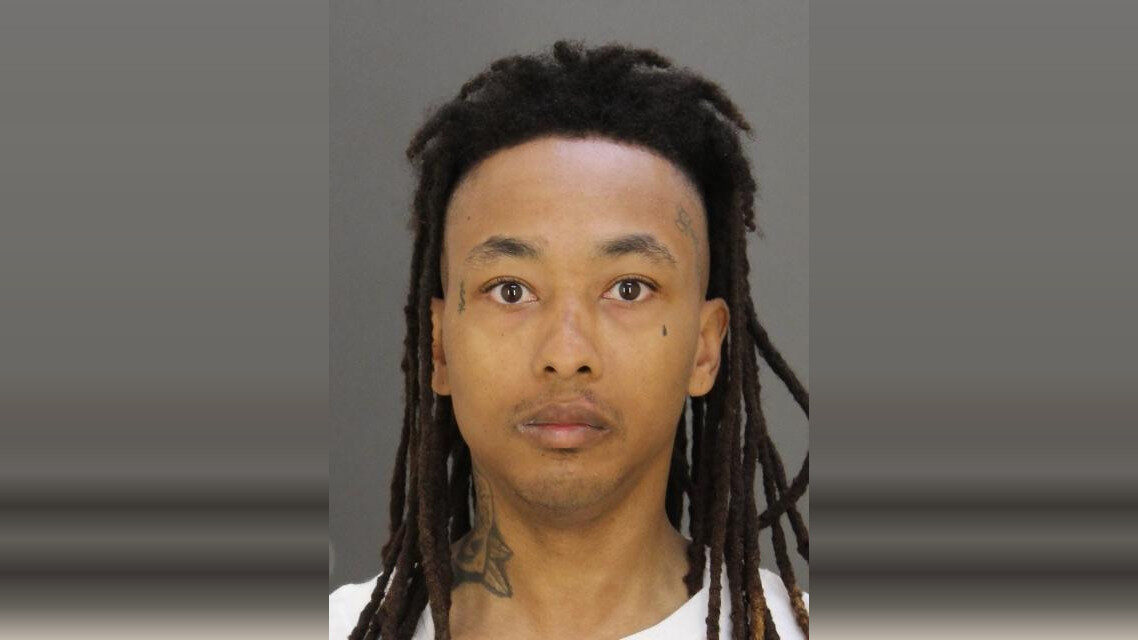 Mugshot-style portrait of a man with long dreadlocks against a plain gray background.