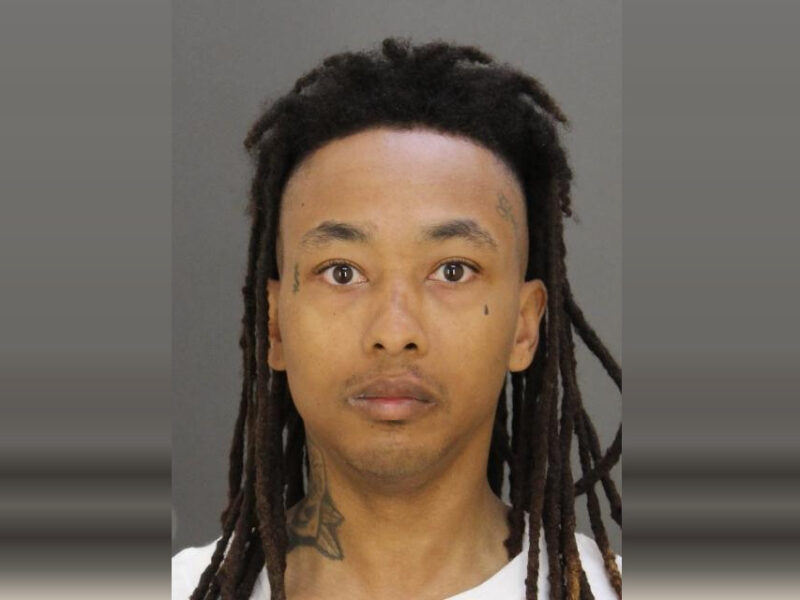 Mugshot-style portrait of a man with long dreadlocks against a plain gray background.