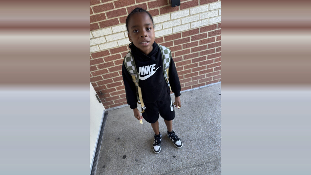 Young boy standing on a sidewalk in front of a brick wall, wearing a black Nike outfit and backpack.