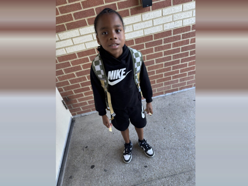 Young boy standing on a sidewalk in front of a brick wall, wearing a black Nike outfit and backpack.