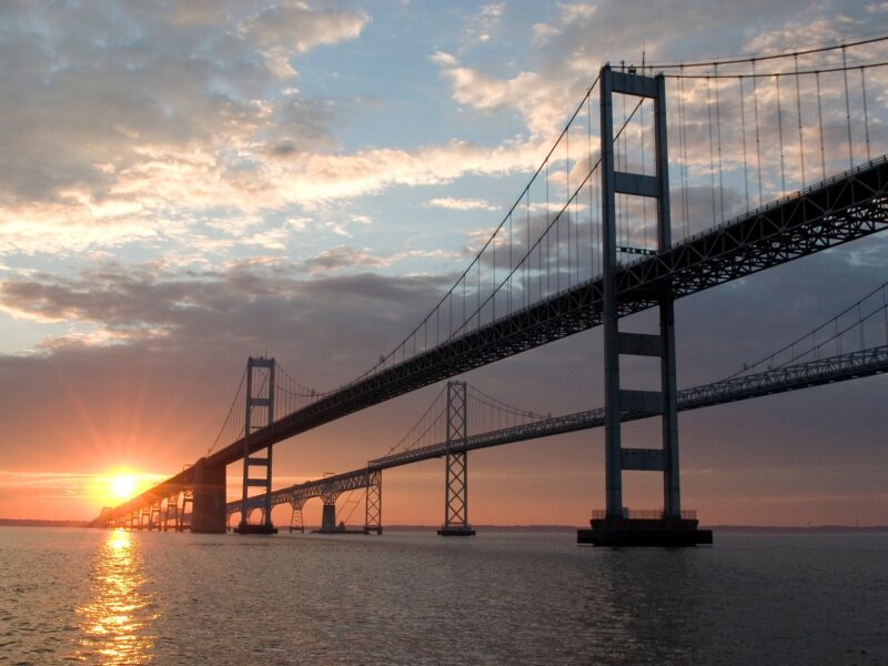 Suspension bridge at sunset over calm water with a glowing orange horizon.