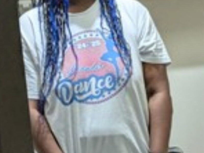 Person with blue braids and glasses standing indoors in a casual outfit.