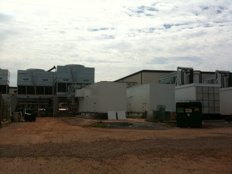 An industrial facility with large white HVAC units and cooling towers. The ground is covered with dirt and gravel, and there is a green dumpster on the right side. The sky is partly cloudy.