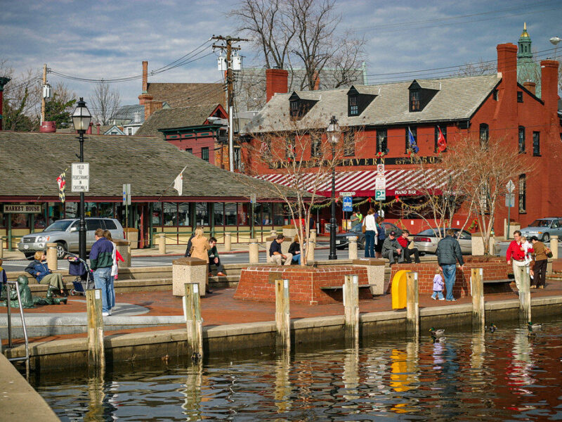 Waterfront harbor scene with people walking near red brick buildings, docks, and a reflective canal.