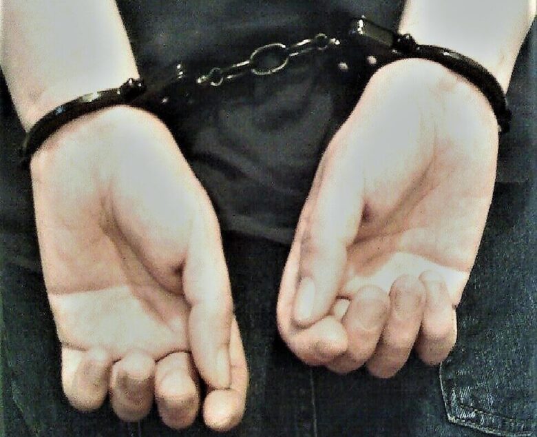 Person's hands cuffed behind their back with black leather restraints.