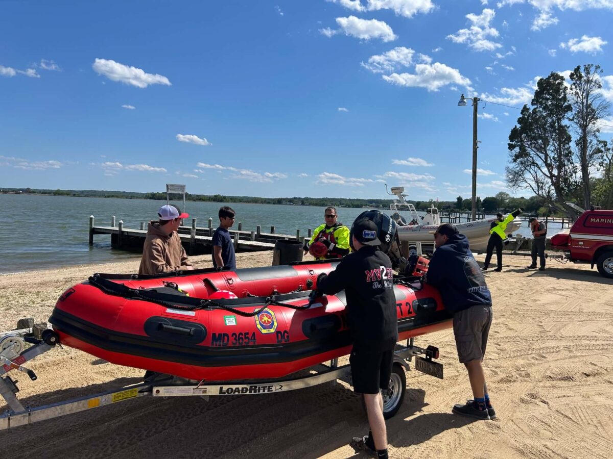 People prepare to launch a red inflatable rescue boat at a sandy lakeside shore.